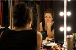 Een vrouw kijkt backstage in een verlichte spiegel in een kleedkamer of theateromgeving. Ze leunt met haar kin op haar hand en kijkt bedachtzaam naar haar spiegelbeeld, omringd door warme lampen en make-upspullen.